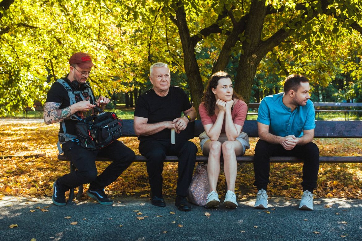 Zespół produkcji filmów reklamowych na ławce w parku. Operator kamery z sprzętem, reżyser i aktorzy podczas przerwy na planie zdjęciowym. Jesienna sceneria z żółtymi liśćmi.
