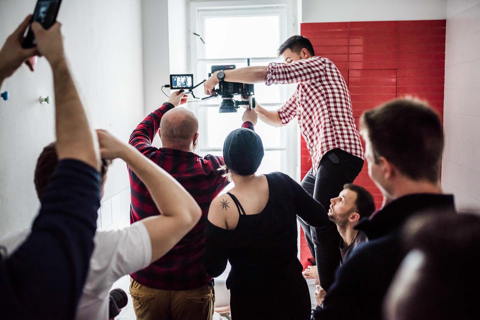 Zespoły na warsztaty 1 Zespół produkcji filmowej pracuje nad nagraniem sceny. Mężczyzna w kratę reguluje kamerę na dźwigu, operatorzy trzymają telefony do dokumentacji, czerwona ściana w tle, światło naturalne z okna.