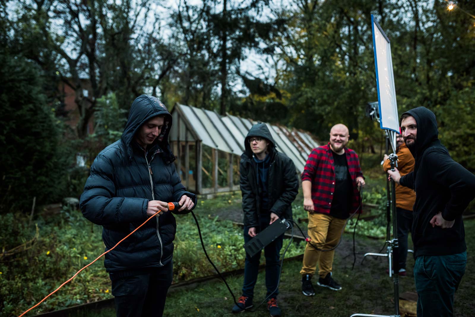 Zespoły na warsztaty 45 Zespół produkcji filmowej na planie w ogrodzie. Mężczyzna w czarnej puchowej kurtce trzyma kabel zasilający, obok niego cztery osoby w zimowych ubraniach przy reflektorze i niebieskim ekranie.