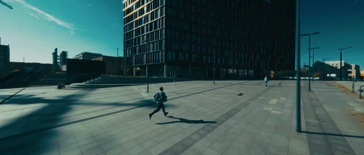 Film promujący wydarzenie Igrzyska Wolności 2 Biegacz w czarnej kurtce przebiega przez przestronny plac miejski otoczony nowoczesną architekturą, błękitne światło, produkcja filmowa.