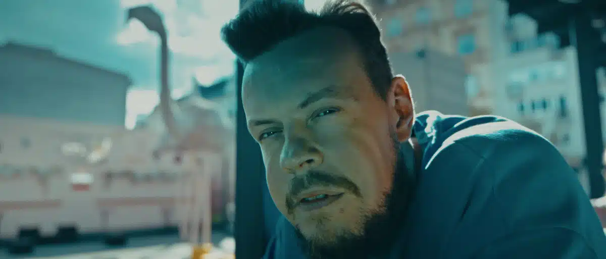 Film promujący wydarzenie Igrzyska Wolności 4 Mężczyzna z brodą w niebieskiej kurtce na dachu z widokiem na miasto, kadr z produkcji filmowej.