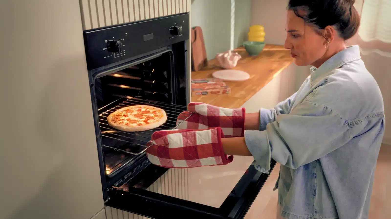 Osoba w rękawicach kuchennych w kratkę wyjmuje pizzę z piekarnika w jasnej kuchni, kadr ze spotu Dr. Oetker Feliciana