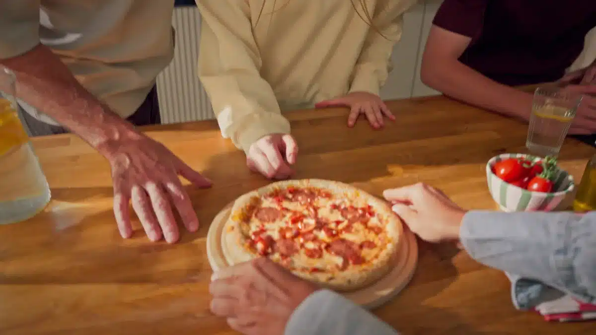 Pizza na desce stawiana na drewnianym stole, dłonie domowników sięgają po kawałki