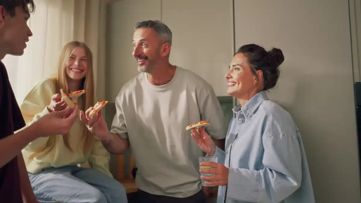 Cztery osoby w kuchni śmieją się z kawałkami pizzy w dłoniach, ciepłe światło